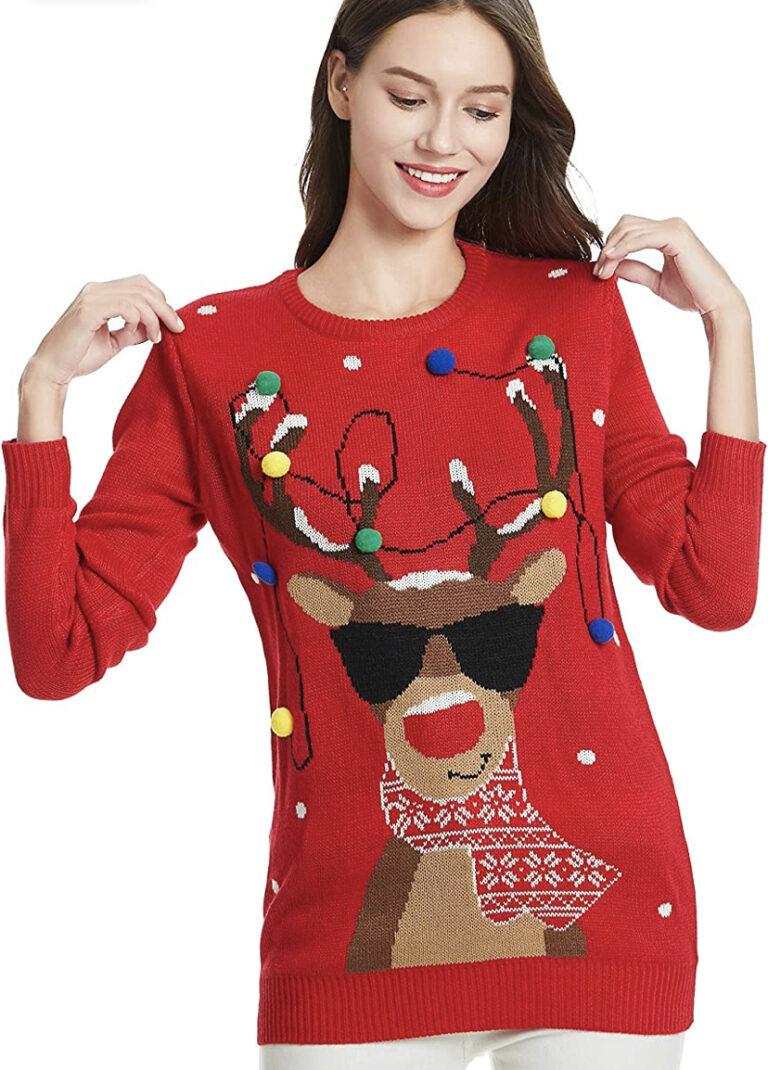 A woman grins in a wild red sweater flaunting a reindeer rocking shades, scarf, pom-poms, and snowflakes—festive chaos perfection.
